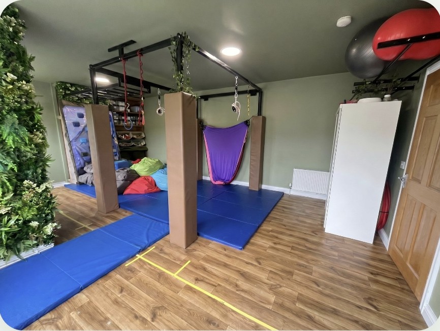 A wider view of the sensory room: a purple therapy hammock on the suspended swing frame, padded posts, blue crash mats, gym balls and a green plant wall